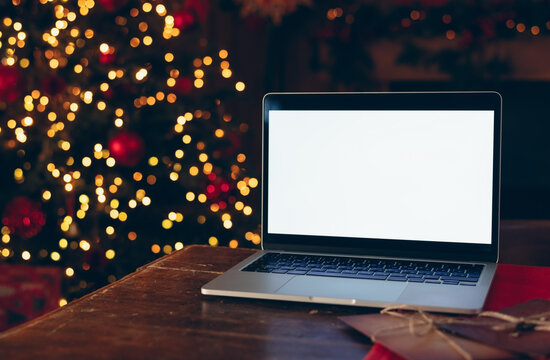 Laptop with a white screen mockup placed on a dark wooden table indoors against a background of a brightly lit, blurred Christmas tree.