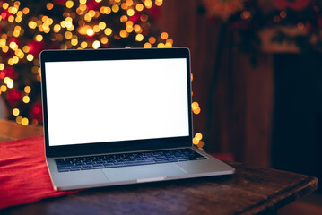 A modern laptop with a blank white screen placed on a wooden table near a blurry background of warm Christmas tree bokeh lights.