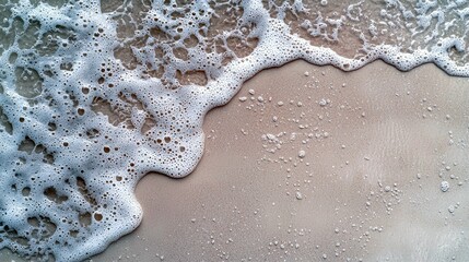 A close-up view of white ocean foam and water gently washing onto a sandy beach, with small bubbles and ripples visible on the wet sand.