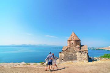 A family admires at the 9th-century architectural historical monument (the Surb Arakelots churche), against the backdrop of the azure lake and hills.