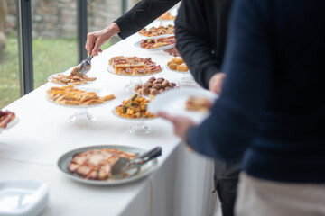 People gathering around a long white table serving delicious food at an outdoor event celebrating a special occasion with a blurred background of lush green trees and a stone wall