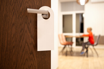 Blank white door hanger mockup hanging on a wooden hotel door handle and blurred silhouette of sitting kid, modern interior with blurred background. Perfect for hospitality branding, hotel signage, an