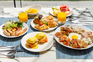 Vibrant breakfast spread on marble table includes eggs Benedict, pastries, bacon, fresh fruit, salads, orange juice. Outdoor dining setting, bright sunlight enhances appetizing display.