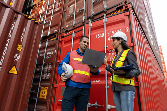 Two serious logistics workers discuss cargo plans near tall red shipping containers, one holding laptop and other hard hat, signifying focused business and import export management at port