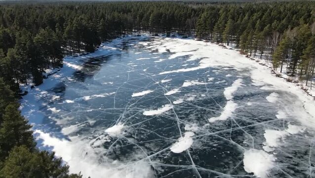 Stunning aerial view of a frozen lake, its surface etched with intricate ice patterns and cracks. A dense evergreen forest frames this serene winter landscape