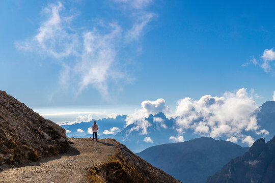 A senior traveler hikes alone along a mountain trail under bright sunlight, surrounded by stunning peaks and clouds, embracing freedom, wellness, and adventure in nature.