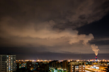 Paesaggio urbano notturno con inceneritore e il fumo che fuoriesce dalle ciminiere. Nuvole scure incombono sul paesaggio urbano illuminato, che comprende un mix di edifici residenziali e industriali