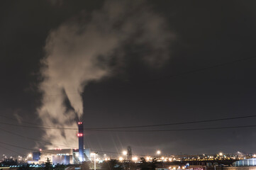 La ciminiera di una centrale elettrica, inceneritore di rifiuti urbani rilascia fumo nel cielo notturno, periferia di una città. L'impianto è illuminato, proiettando luce sull'ambiente circostante.