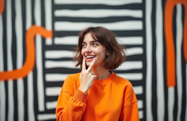 Young smiling woman with short brown hair wears orange sweatshirt. She gestures with two fingers towards camera. Her playful expression matches bold striped background. Outdoor urban setting.