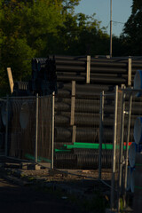 A collection of stacked black cylinders behind a fence, indicating construction activity. Green surface visible in foreground.