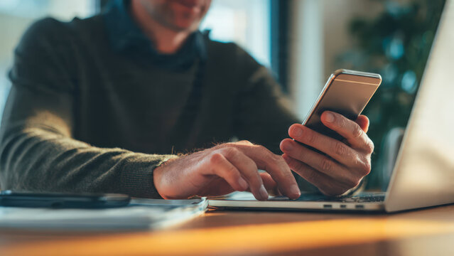 Close-up of a man using a laptop while holding a smartphone, representing multitasking, digital communication, remote work, and modern productivity.