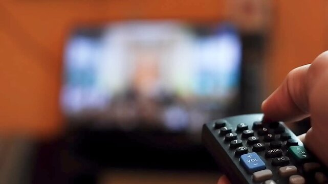 A close-up hand holding a TV remote control to switch off the power while pointing toward a blurred television screen in a dimly lit room