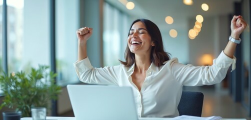Woman celebrates success at laptop. She smiles widely with arms raised in office. Financial freedom or crypto trading gains achieved. Happiness and winning at work.