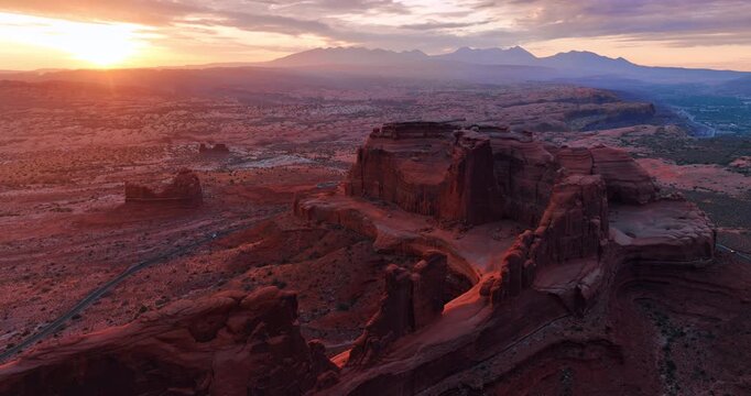 Wonderful rocks with steep walls from drone footage. Few cars go around the rock in the Arches National Park in Utah, USA. Beautiful setting sun in the sky over the scenery.