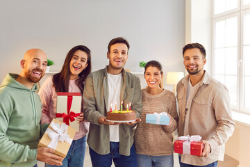 Portrait of a smiling young happy man holding birthday cake with candles standing with friends holding present gift boxes indoors and looking cheerful at camera. Birthday party concept.