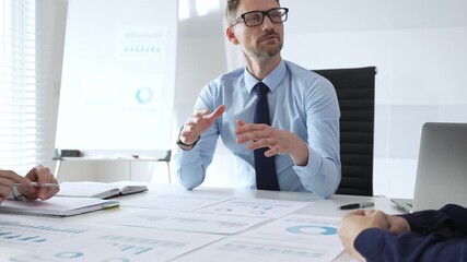 Confident businessman in glasses leads a corporate meeting, reviewing financial charts, explaining data insights, and discussing strategic growth opportunities with colleagues