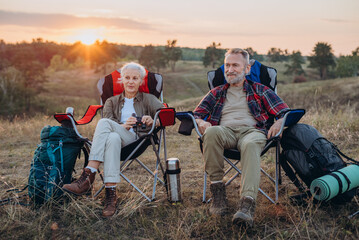 Senior couple sits on camping chairs watching setting sun over quiet valley