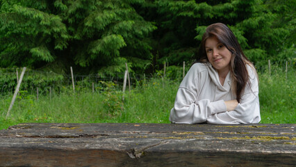 Smiling Young Woman Leaning on a Rustic Wooden Table in a Green Forest or Park Setting, Looking at the Camera.