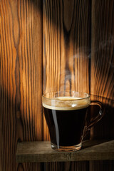 Ray of light falls on transparent cup of coffee standing on shelf against wooden background