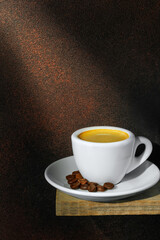 Ray of light falls on small white cup espresso with saucer and coffee beans standing on board against brown background