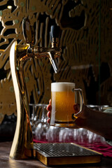 A hand holds a frosty mug filled with golden beer while a shiny tap pours the last drops. The bar has a cozy atmosphere, illuminated gently by warm lights, perfect for relaxation