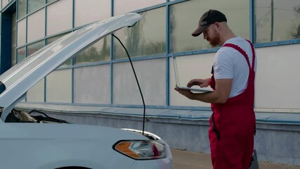 Technician in red overalls using diagnostic computer to troubleshoot engine issues, standing beside open hood of vehicle in a well-lit automotive workshop environment