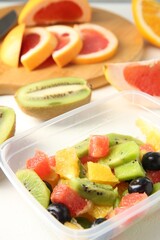 Tasty fruit salad in plastic container and ingredients on white table, closeup. Healthy snack