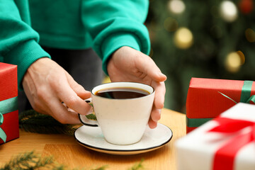 Woman with cup of aromatic coffee and gift boxes at wooden table against blurred lights, closeup. Christmas greeting card