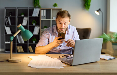 Business man accountant with phone and laptop feels anxiety among fiancial documents. In office, employee reading bad news message, driving work stress, confusion, frustration and burnout. 