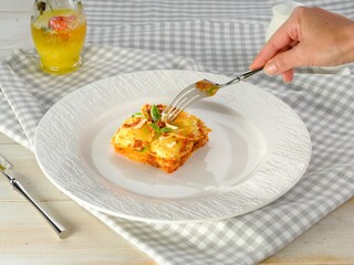 A hand gracefully lifts a forkful of cheesy pasta from a neatly arranged dish. A refreshing drink sits beside, enhancing the dining experience in a cozy setting