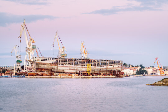 Large industrial crane and ship hull under construction at a shipyard near the water, illuminated by soft evening light. Maritime industry and engineering concept. - Powered by Adobe