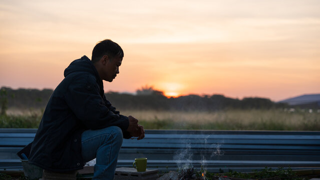Young Asian man sitting by a campfire at sunrise, feeling calm and thoughtful. - Powered by Adobe