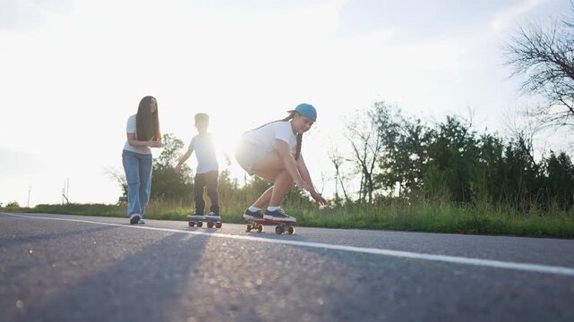 A girl on a skateboard skates with another girl, helping a boy on a skateboard. The boy balances as the girl guides him with her skateboard on the open road.