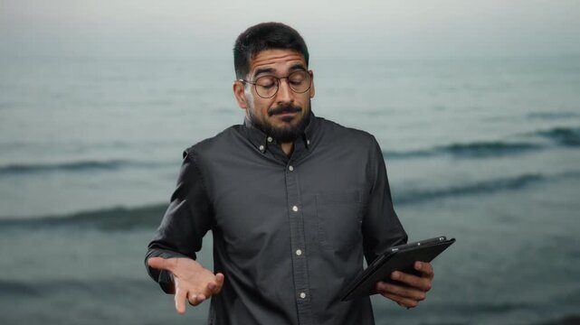 Man standing on beach holding tablet, ocean in background suggesting peaceful outdoor setting, young male appearing thoughtful and engaged, isolated at beautiful seaside location.