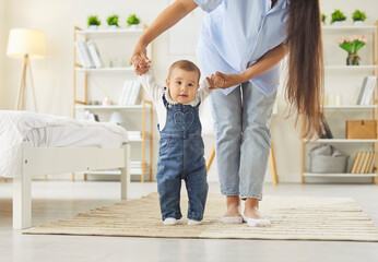 A mother supports her baby learning to walk on a soft rug in a bright bedroom, holding both hands...