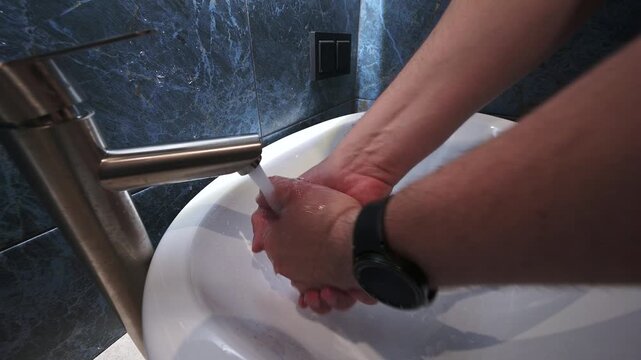 A person thoroughly washes their hands in a modern, clean bathroom sink with dark marble tiles and a sleek chrome faucet, emphasizing hygiene - Powered by Adobe