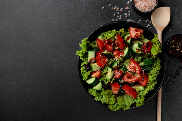 Vegetable salad of tomatoes, cucumbers, onions, lettuce dressed with aromatic dressing in a black plate on a gray background