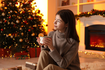 Beautiful woman with cup of tasty cocoa near Christmas tree and fireplace at home