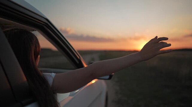 Adventure road under sunset sky. Girl lifts hand from moving car. Nature around road reflects light. Freedom glows on girl. Car and travel scene summer wind. Hand meets sunset moment on road again