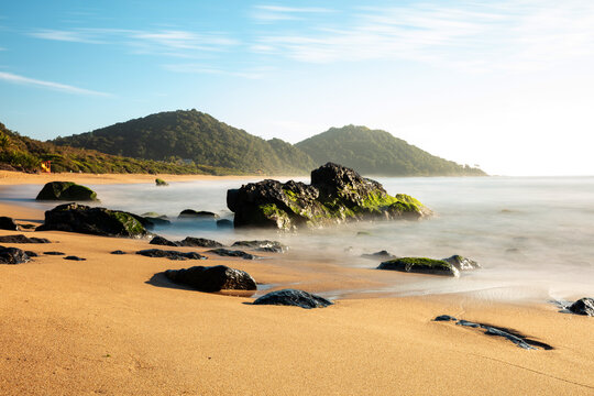 Ocean waves crashing against the rocks of Balne&aacute;rio Cambori&uacute; beach, the sun reflecting on the water, a long exposure at sunrise.