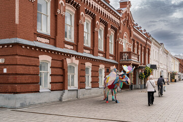 Street in Ulyanovsk with a red brick building and a colorful moose sculpture. People walk along the sidewalk under an overcast sky.