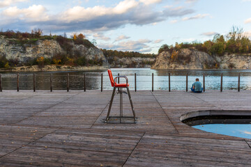 Fototapeta premium Empty red lifeguard chair stands on a wooden lakeside platform overlooking calm water and rocky cliffs. Soft afternoon light and still reflections create a minimal yet atmospheric scene of solitude an