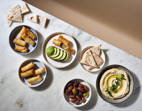 Overhead shot showcasing a vibrant spread of spring rolls, pita bread, hummus, avocado, and olives on a marble background, perfect for food blogging or recipe sharing. - Powered by Adobe