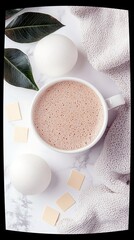 A top-down view of a white mug filled with frothy hot chocolate, surrounded by bath bombs, green leaves, and a textured blanket on a marble surface.