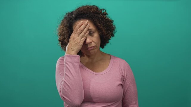 Woman wearing pink shirt places hand on forehead, closing eyes in frustration in studio with teal background; fatigue.
