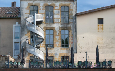 A restaurant terrace in Mont de Marsan features tables with chairs and a unique spiral staircase on the building. The inviting space is perfect for enjoying meals outdoors.