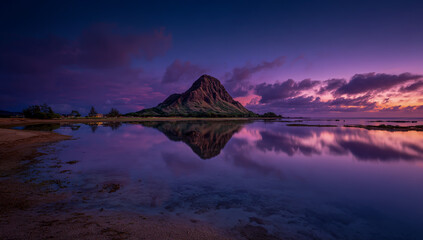Dramatic purple and pink sunset sky reflects on calm ocean water with a prominent rock formation
