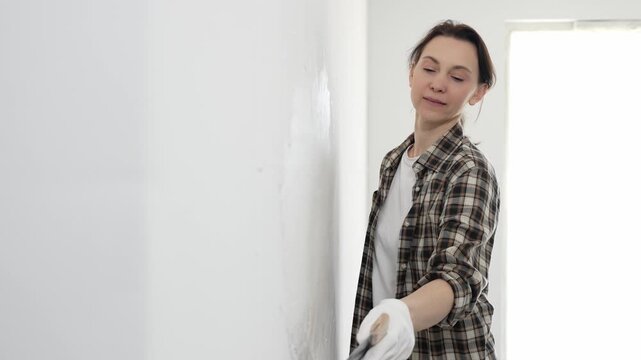 Female construction professional wearing checkered shirt is smoothing white wall surface, applying putty with professional spatula technique, showcasing skilled interior renovation work