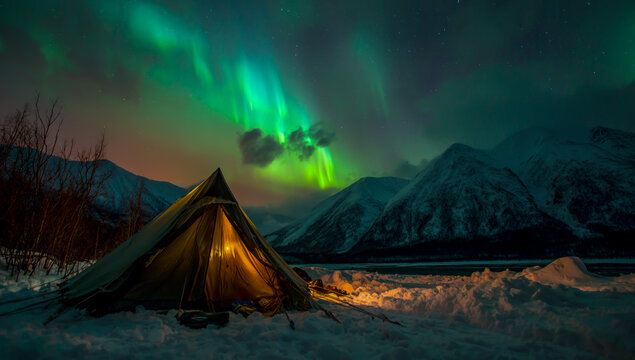 Cozy illuminated tent under vibrant green aurora borealis dancing over snow covered mountains