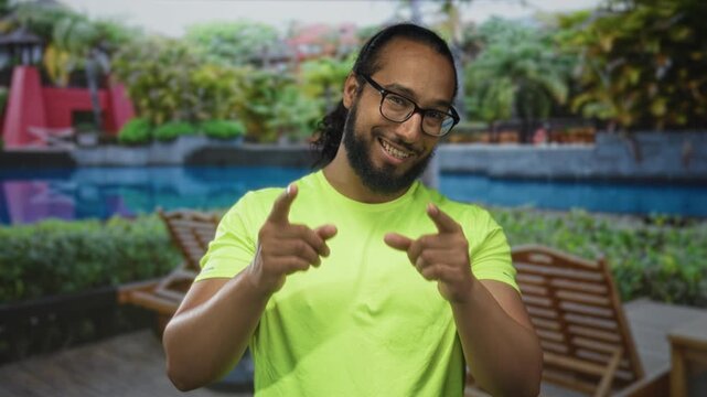 Man points fingers at camera beside hotel pool and wooden lounger wearing bright green shirt; friendly confidence.